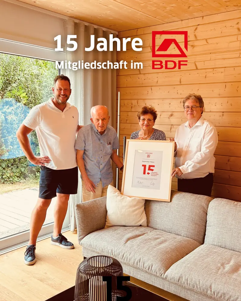 Gruppenfoto in einem Stommel-Haus-Innenraum mit Holzwand und großer Fensterfront. Vier Personen stehen gemeinsam vor einer eingerahmten Urkunde zur 15-jährigen Mitgliedschaft im Bundesverband Deutscher Fertigbau (BDF). Die Urkunde wurde bei einem Besuch des BDF-Geschäftsführers in der Stommel Haus Manufaktur in Eischeid überreicht. Im Bild sichtbar sind außerdem das BDF-Logo sowie der Schriftzug „15 Jahre Mitgliedschaft im BDF“.