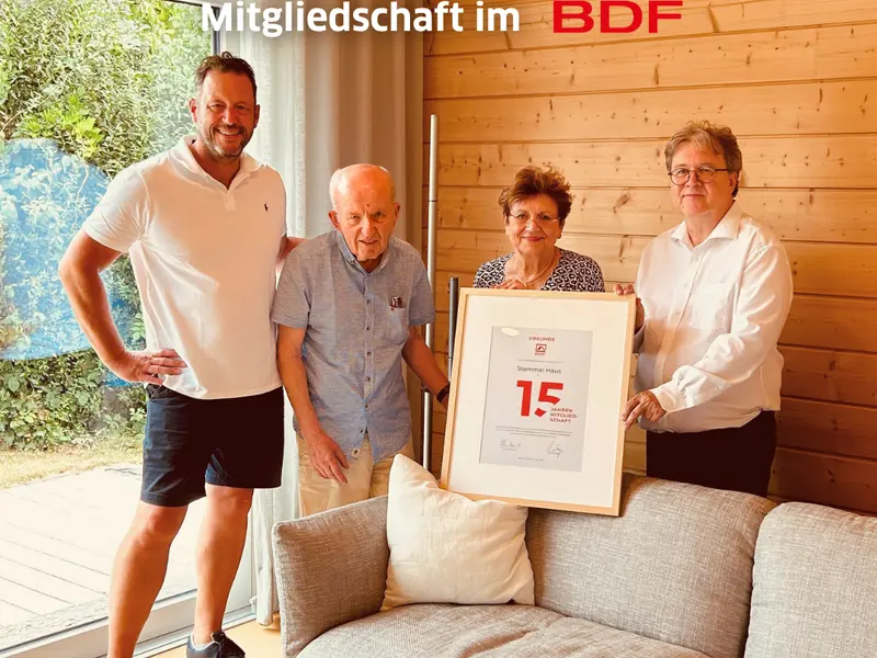 Gruppenfoto in einem Stommel-Haus-Innenraum mit Holzwand und großer Fensterfront. Vier Personen stehen gemeinsam vor einer eingerahmten Urkunde zur 15-jährigen Mitgliedschaft im Bundesverband Deutscher Fertigbau (BDF). Die Urkunde wurde bei einem Besuch des BDF-Geschäftsführers in der Stommel Haus Manufaktur in Eischeid überreicht. Im Bild sichtbar sind außerdem das BDF-Logo sowie der Schriftzug „15 Jahre Mitgliedschaft im BDF“.