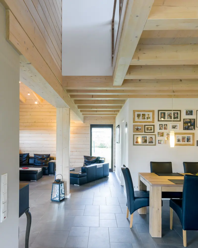Offener Wohn- und Essbereich in einem Massivholzhaus mit sichtbarer Holzdecke, Holzbalken, Sitzbereich, Esstisch und hellen Wandflächen.