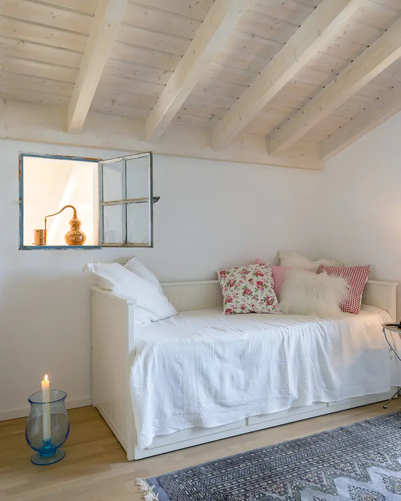 Heller Schlafraum mit sichtbarer Massivholzdecke, Tagesbett, dekorativen Textilien und wohnlicher Atmosphäre.