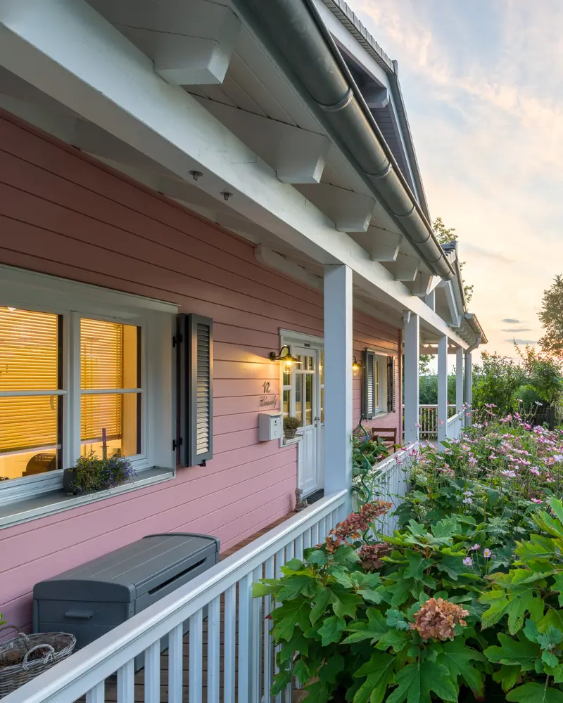 Nahaufnahme einer überdachten Veranda mit rosa Holzfassade, weißen Stützen, grauen Fensterläden, warm beleuchteten Fenstern und dichtem Gartenbewuchs.