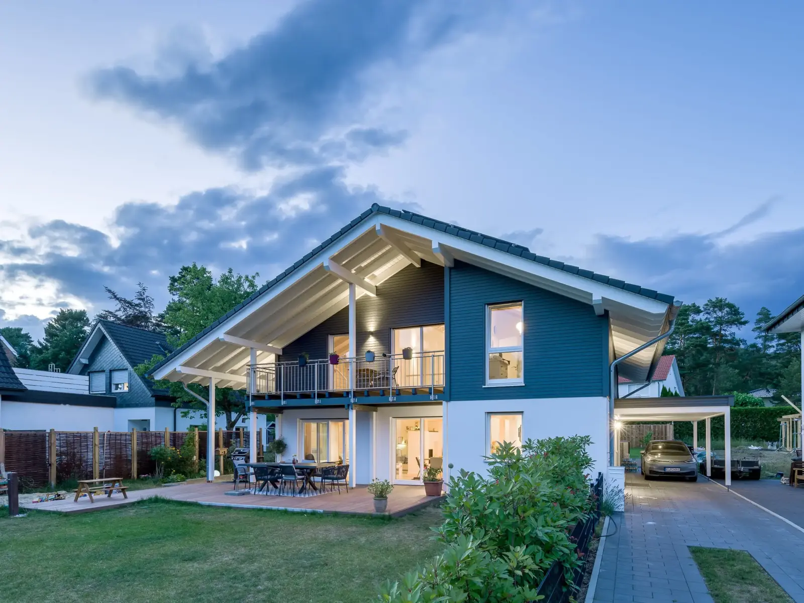 Anthrazitfarbenes Landhaus von Stommel Haus mit Carport, Balkon und beleuchteten Fenstern in der Abenddämmerung.