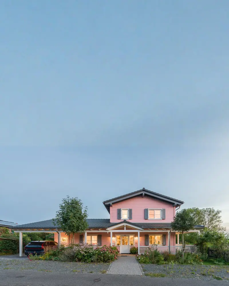 Frontansicht eines rosa Massivholzhauses mit symmetrischem Eingangsbereich, weißer Veranda, grauen Fensterläden und bepflanztem Vorgarten.