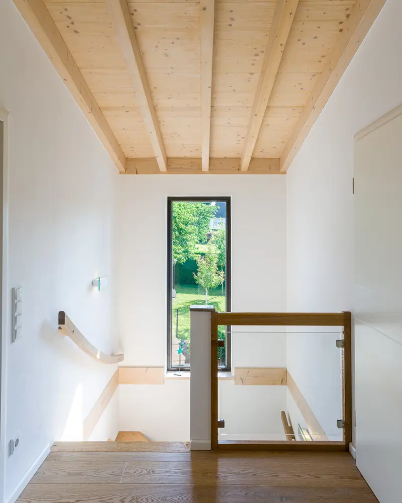 Heller Treppenbereich in einem Massivholzhaus mit sichtbarer Holzdecke, Holzboden, Glasgeländer und schmalem Fenster zum Garten.