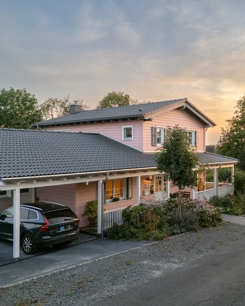 Rosa Holzhaus mit überdachtem Carport, Veranda, grauem Ziegeldach, Gartenbepflanzung und warmem Abendlicht am Eingang.