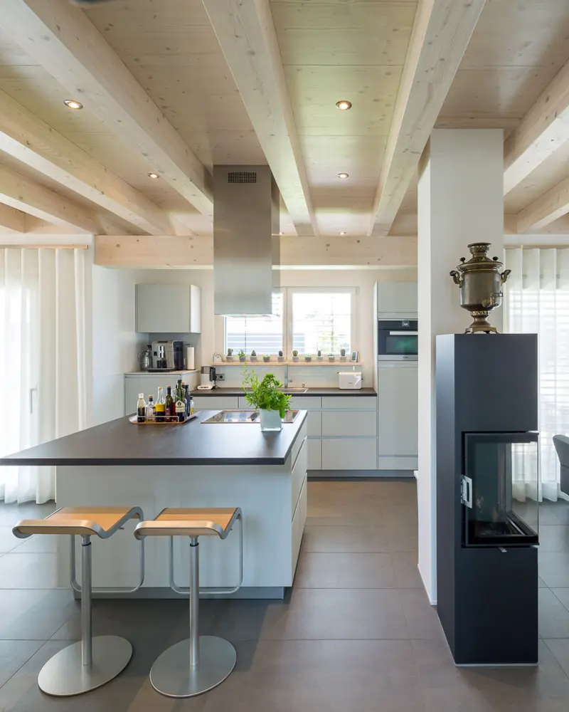 Moderne Wohnküche mit Kochinsel, sichtbaren Holzbalken und hochwertiger Innenausstattung in einem individuellen Holzhaus.
