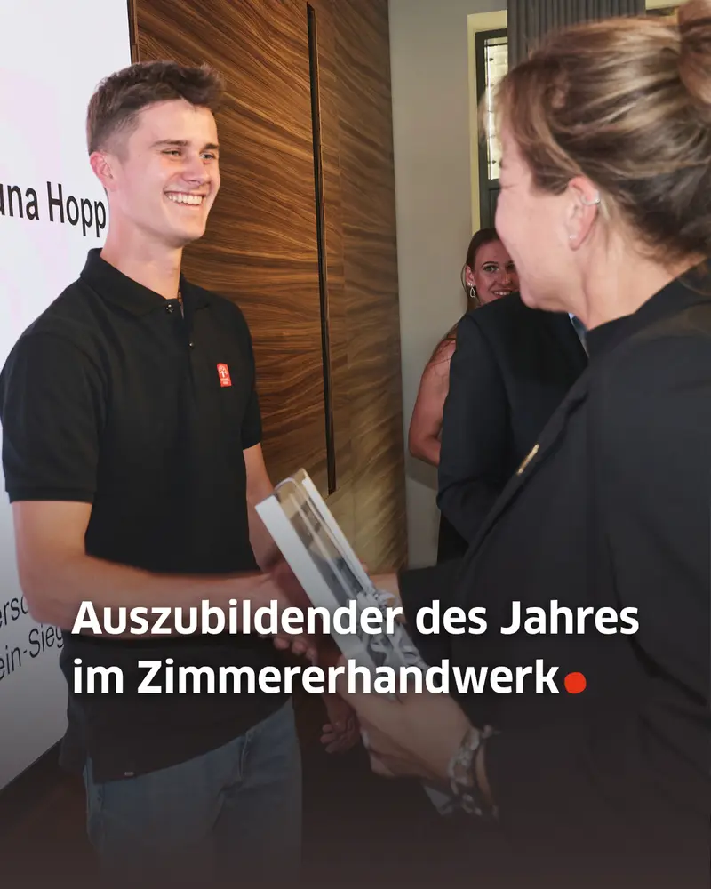 Eine ausgezeichnete Person in schwarzem Poloshirt nimmt lächelnd eine gerahmte Urkunde entgegen. Im Vordergrund ist die überreichende Person von hinten zu sehen. Auf dem Bild steht: „Auszubildender des Jahres im Zimmererhandwerk.“