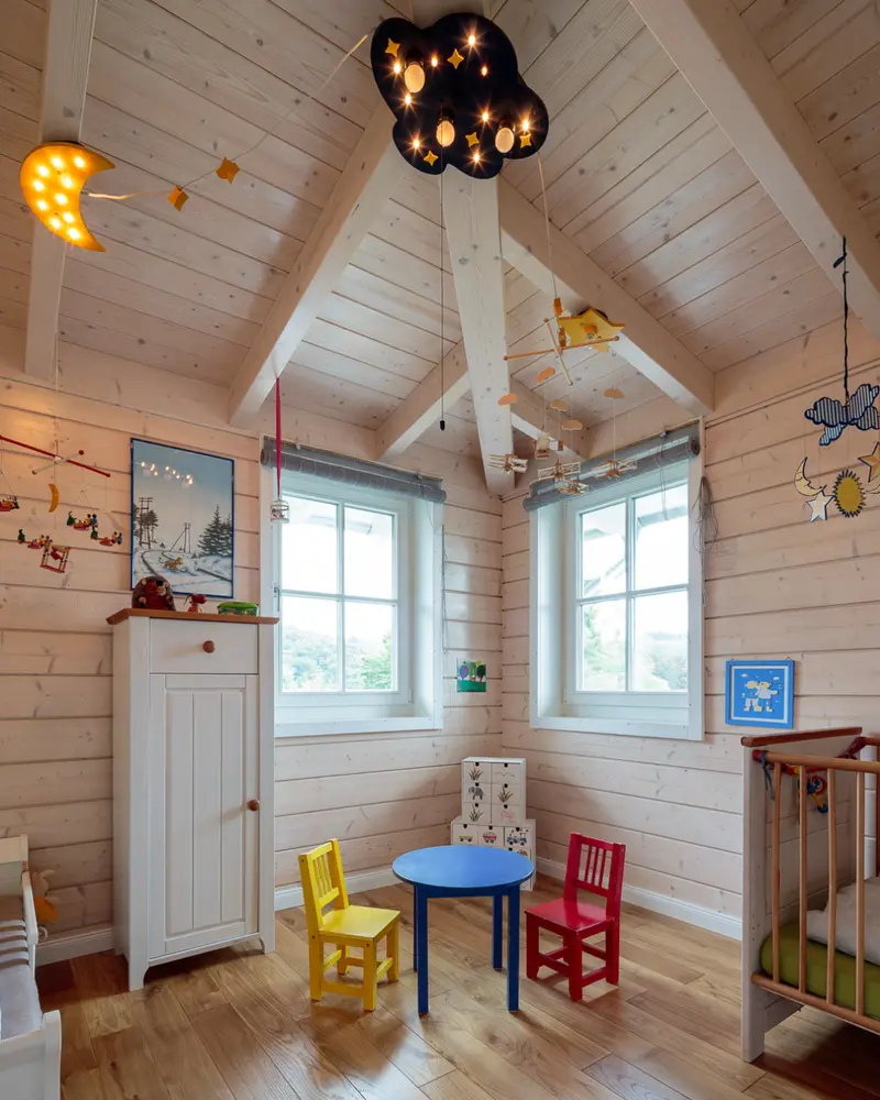 Kinderzimmer im Holzhaus mit heller Holzverschalung, sichtbaren Dachbalken, zwei Sprossenfenstern, bunten Stühlen und Spielmöbeln.