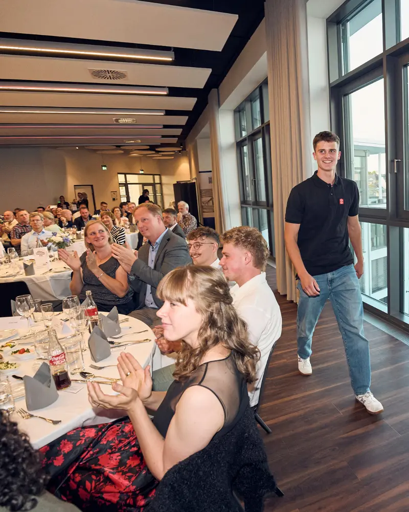 Eine Person in schwarzem Poloshirt geht am Rand des Veranstaltungssaals entlang. An den gedeckten Tischen sitzen zahlreiche Gäste und applaudieren. Große Fenster und eine Holzdecke prägen den Raum.
