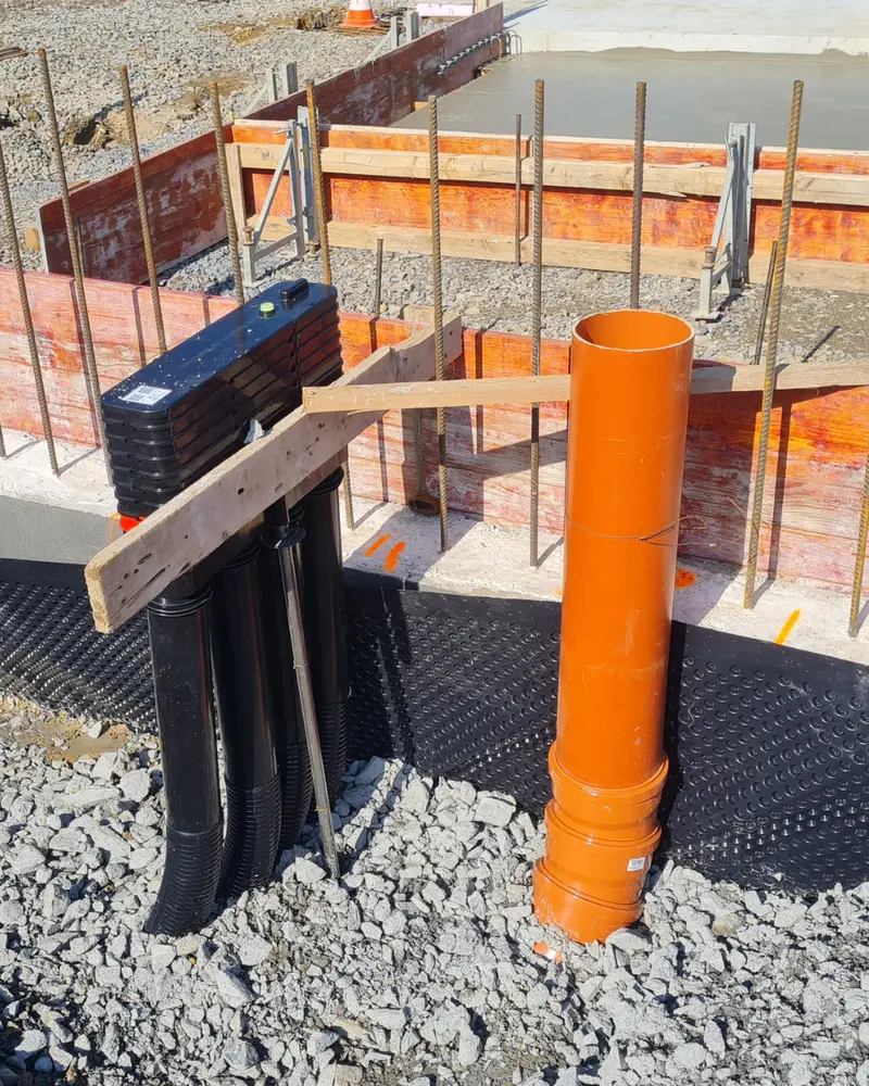 Baustelle mit vorbereiteten Leitungsrohren, orangefarbenem Kanalrohr, Schalung und Bewehrung vor dem Betonieren der Bodenplatte.