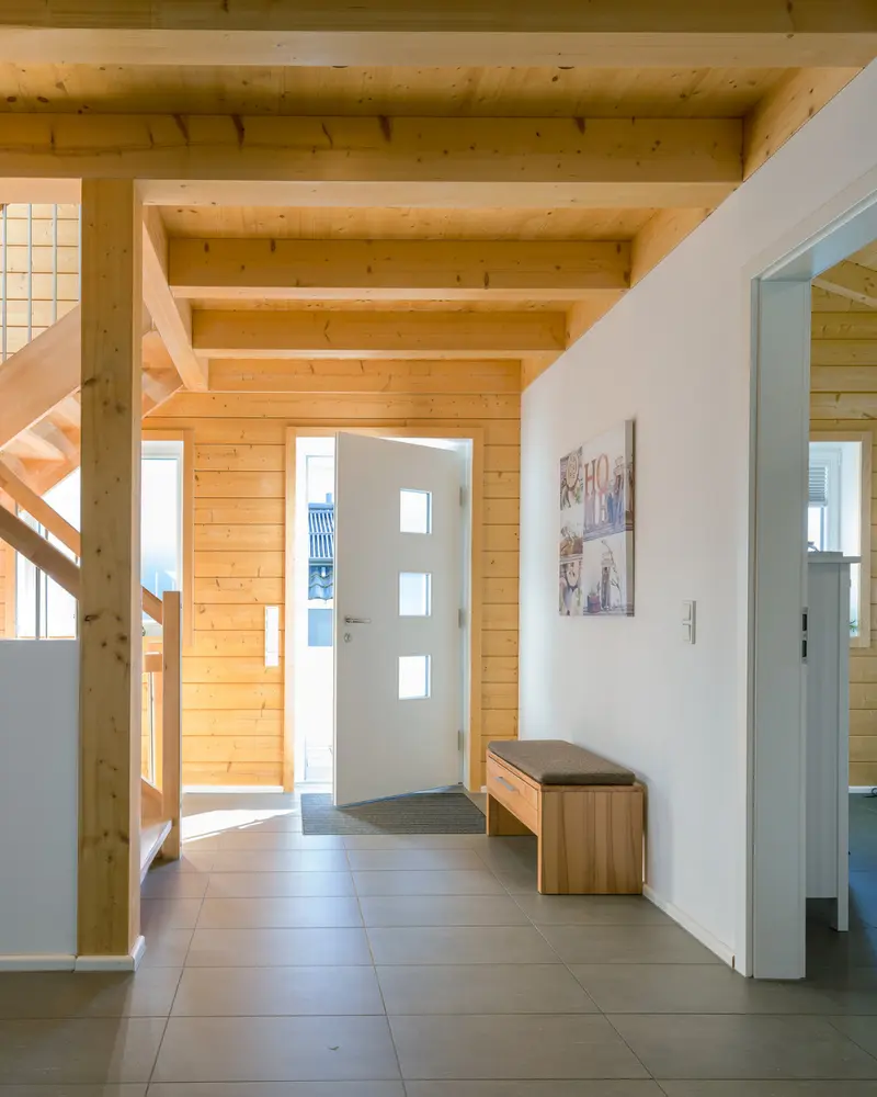 Heller Eingangsbereich eines Holzhauses mit sichtbaren Deckenbalken, Holzwänden, Treppe, grauen Bodenfliesen und geöffneter Haustür.