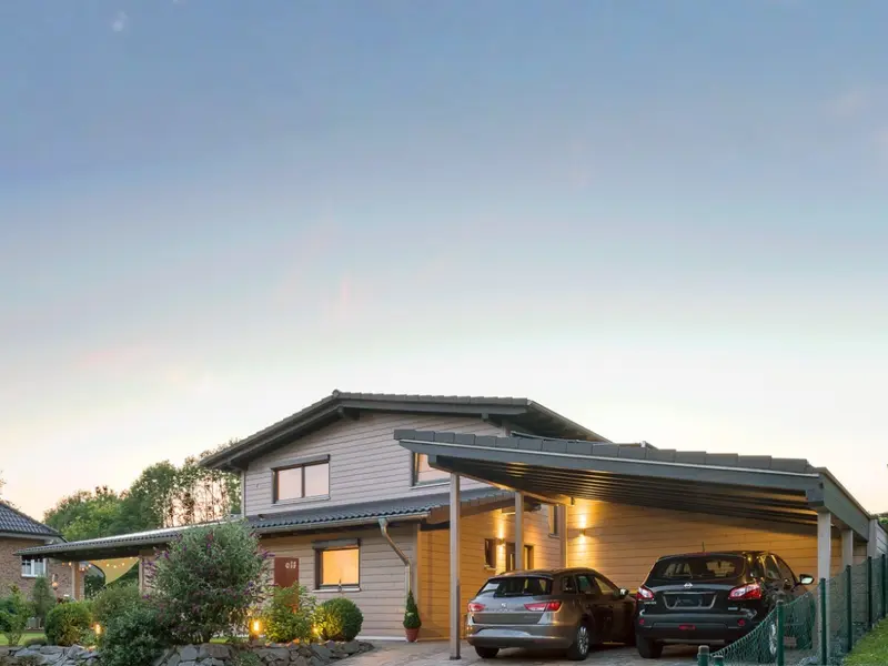 Modernes Massivholzhaus mit heller Holzfassade, beleuchteter Einfahrt, Carport und Garten bei einsetzender Dämmerung.