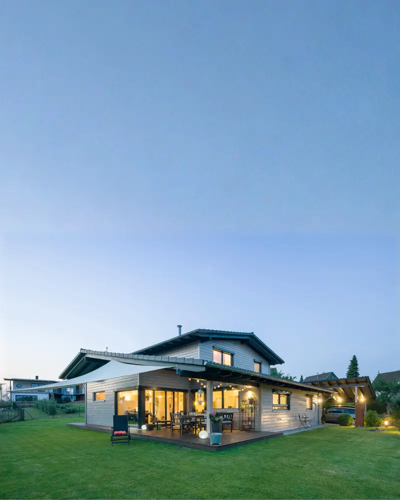 Freistehendes Massivholzhaus mit großer Terrasse, Gartenfläche, Holzfassade und warm beleuchteten Wohnräumen am Abend.