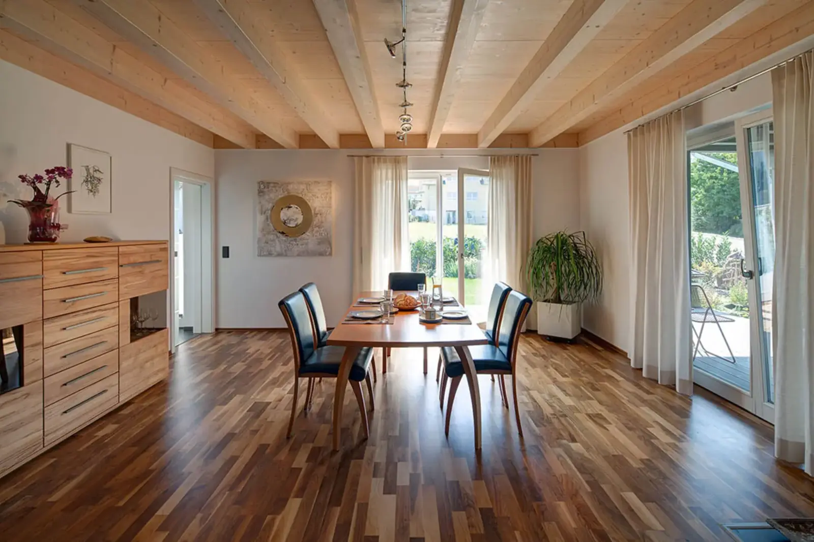 Der Innenraum eines schlüsselfertigen Holzhauses von Stommel Haus. Zu sehen ist ein großes Esszimmer, ein Tisch und Stühle. Ein Regal steht an der Wand, an den anderen Wenster sind große schöne Fenster zu sehen. Der Raum bietet viel Platz.