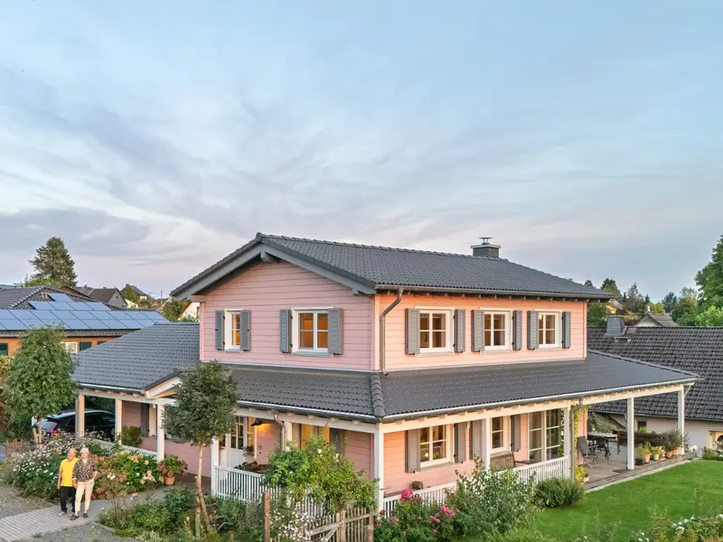 Rosa Holzhaus im American Style mit umlaufender Veranda, grauem Dach, weißem Geländer, Garten und zwei Personen am Eingangsweg.