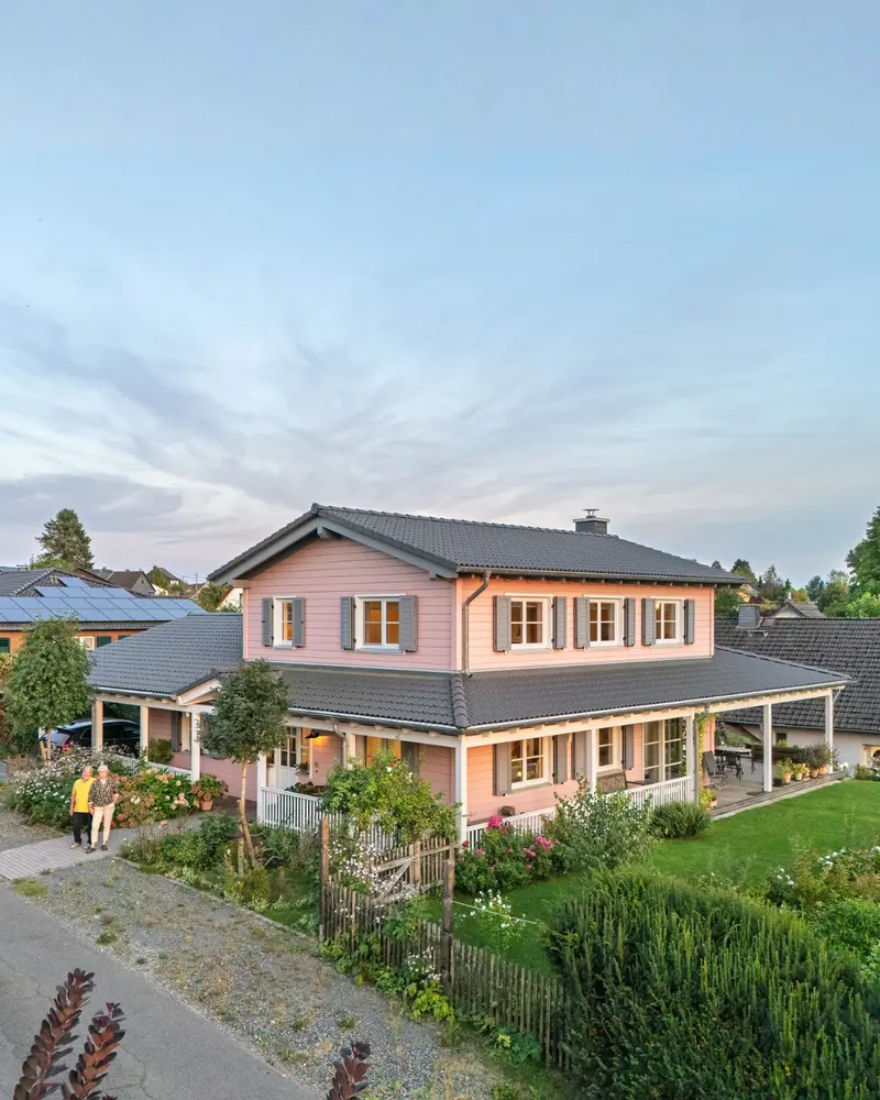 Rosa Holzhaus im American Style mit umlaufender Veranda, grauem Dach, weißem Geländer, Garten und zwei Personen am Eingangsweg.