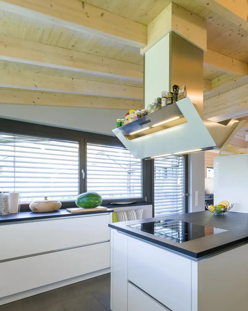 Helle Küche in einem Holzhaus mit Kochfeld, Dunstabzug, großen Fenstern und sichtbarer Holzdecke als Beispiel für vorausschauend geplante Anschlüsse.
