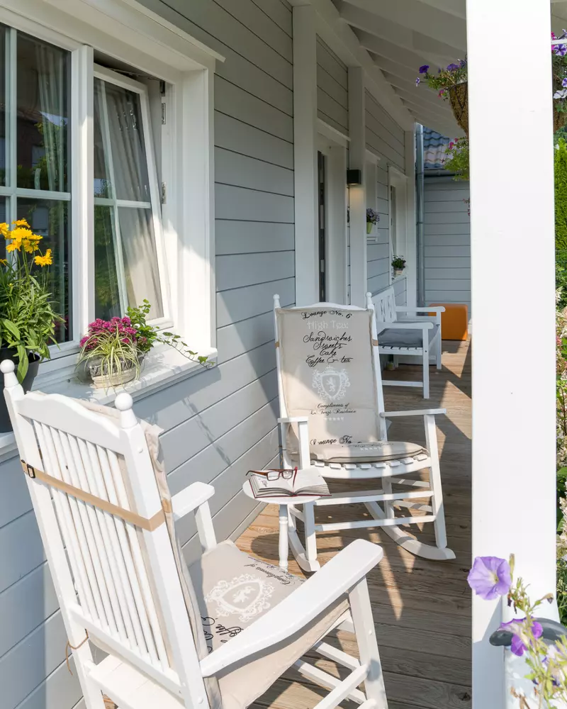 Veranda mit weißen Schaukelstühlen, Holzbohlenboden und dekorierten Fenstern mit Blumenkästen