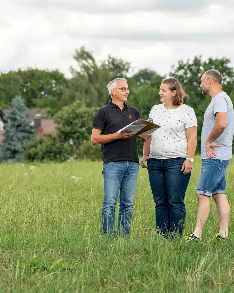 Drei Personen stehen auf einer Wiese und sprechen über Unterlagen zur Hausplanung; im Hintergrund sind Bäume und eine ländliche Umgebung zu sehen.