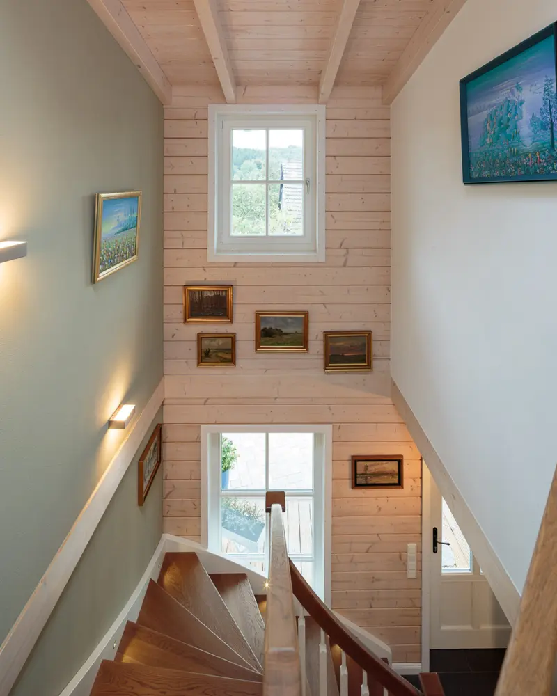 Innenansicht eines Holzhauses mit Treppenaufgang, heller Holzverschalung, sichtbaren Deckenbalken, kleinen Bildern und weißen Fenstern.