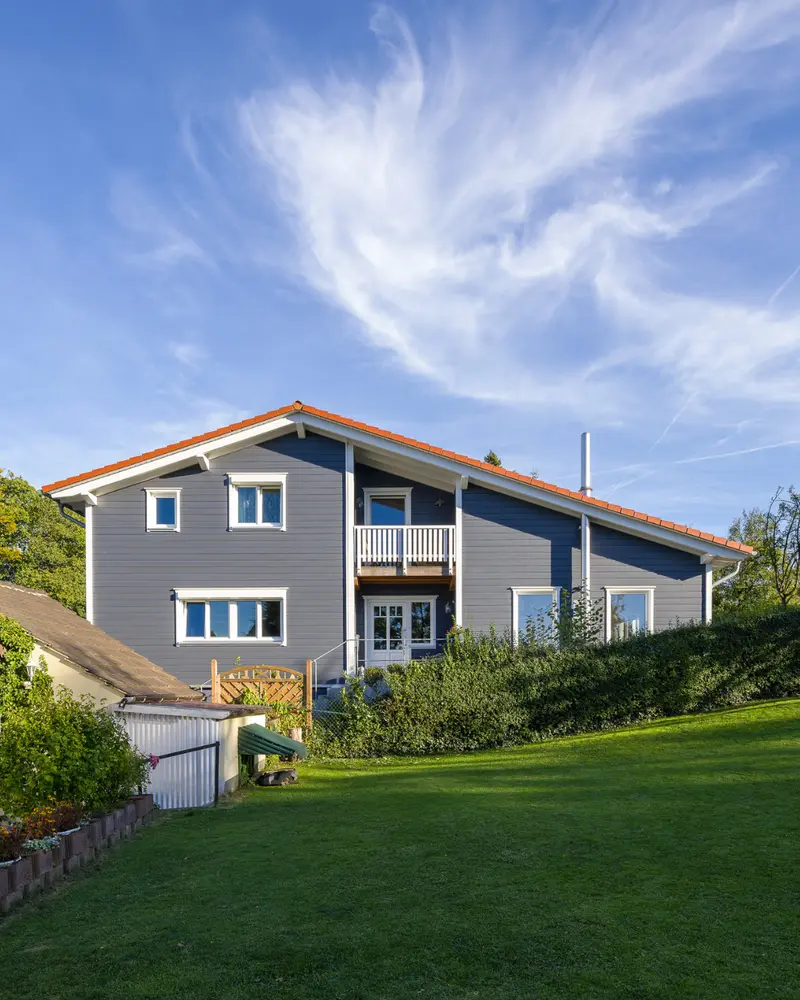 Individuell geplantes Holzhaus von Stommel Haus auf leicht geneigtem Grundstück mit großer Rasenfläche, gepflegter Bepflanzung und markanter Holzfassade unter blauem Himmel.