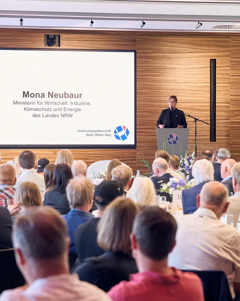 Blick über das sitzende Publikum auf eine Person am Rednerpult. Auf der Leinwand steht „Mona Neubaur, Ministerin für Wirtschaft, Industrie, Klimaschutz und Energie des Landes NRW“. Darunter sind Name und Logo der Kreishandwerkerschaft Bonn•Rhein-Sieg zu sehen.