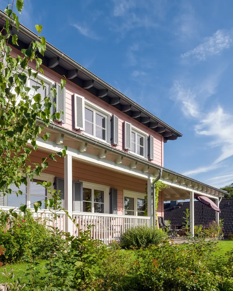Seitliche Ansicht eines rosa Holzhauses mit langer Veranda, weißen Geländern, grauen Fensterläden, großen Fenstern und gepflegtem Garten.