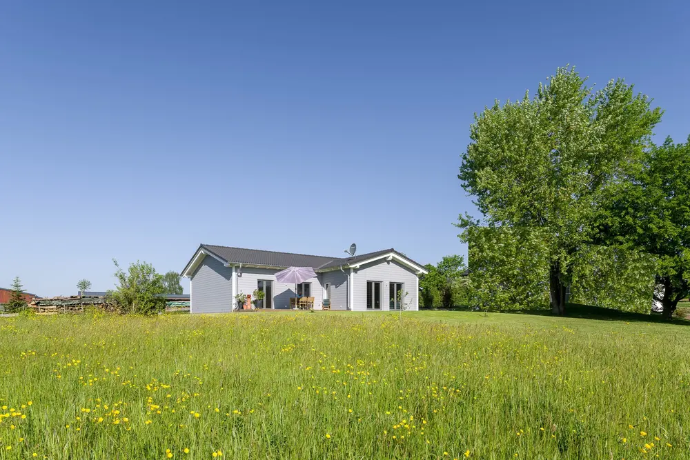 Ein ebenerdiges kleines Haus für Senioren von Stommel Haus mit heller Holzverkleidung und überdachter Terrasse, im Vordergrund eine blühende Wiese.