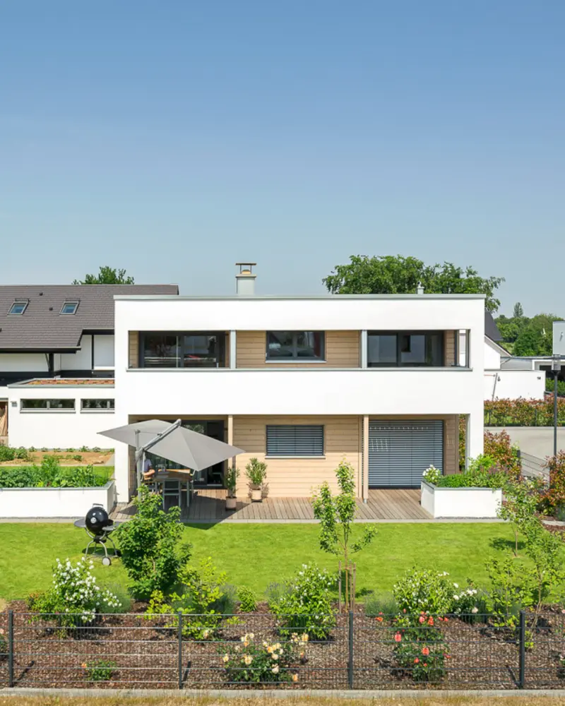 Rückansicht eines modernen Holzhauses mit großer Terrasse, gepflegtem Garten, Bepflanzung und großzügigen Freiflächen.