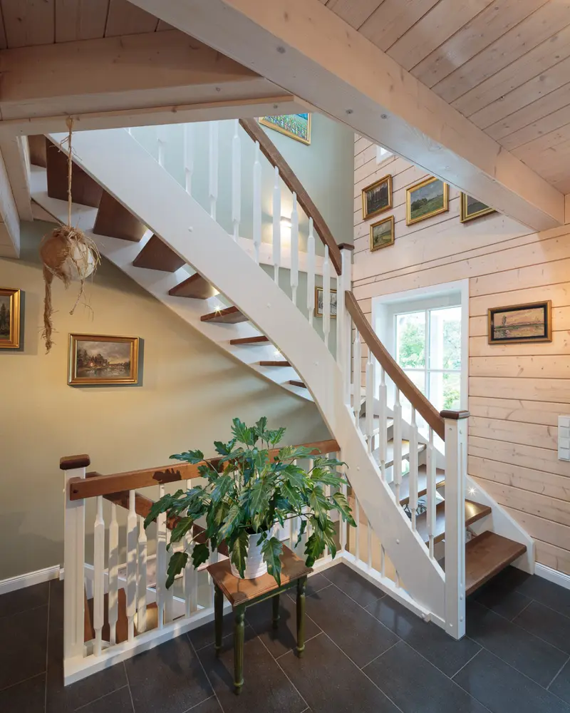 Offener Treppenbereich in einem Holzhaus mit Holzbalkendecke, heller Wandverkleidung, weißen Geländern und dunklem Fliesenboden.