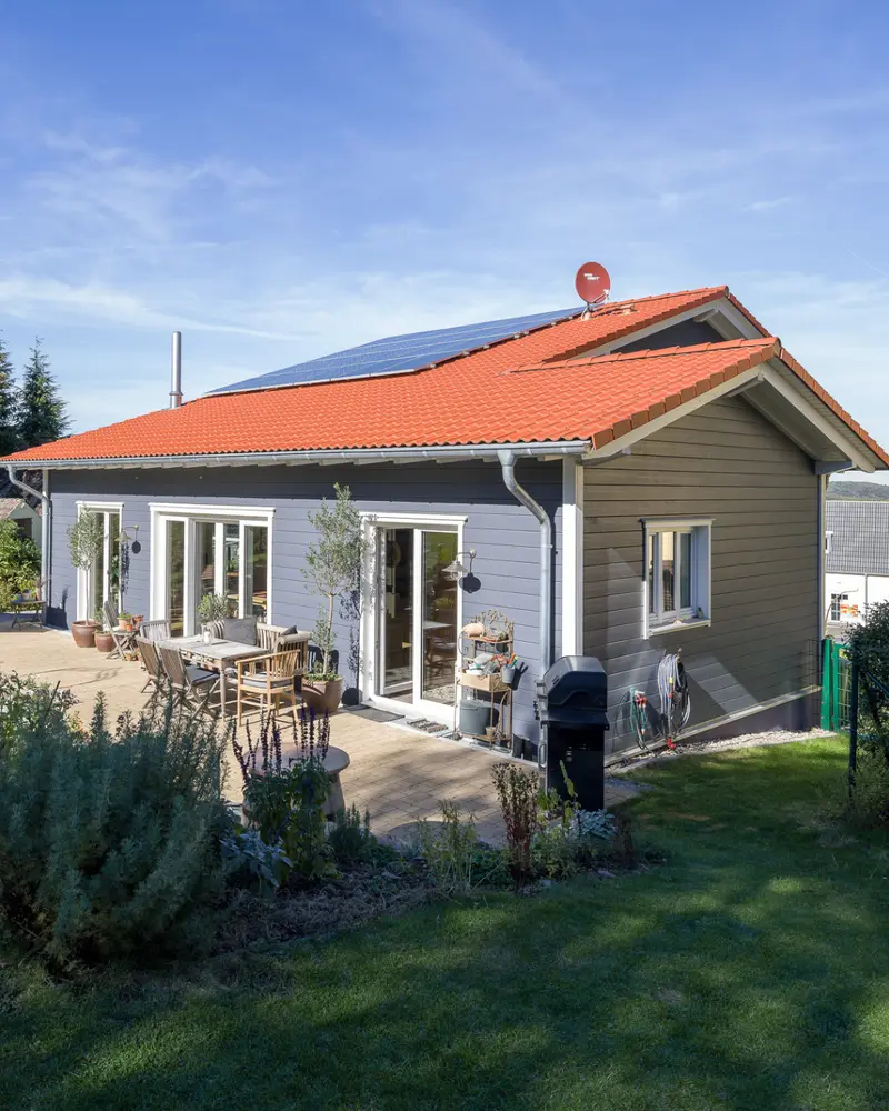 Modernes Holzhaus mit großzügiger Terrasse, großen Fensterelementen, Photovoltaikanlage auf dem Dach und naturnaher Gartengestaltung.