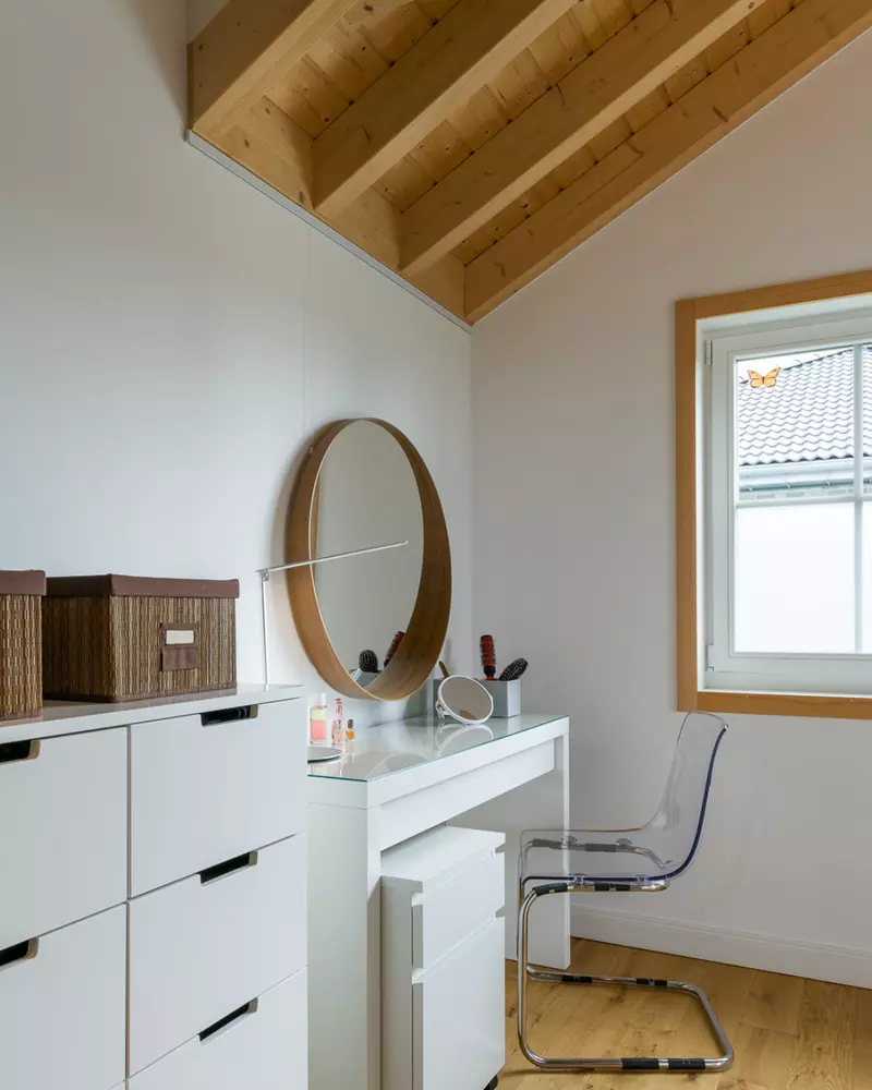 Heller Schminkplatz mit rundem Holzspiegel, weißem Tisch und Schubladen sowie Fenster mit Holzrahmen und Blick nach draußen