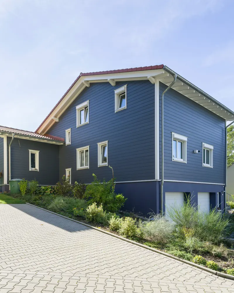 Holzhaus mit dunkler Fassadenverkleidung auf einem Hanggrundstück mit gepflasterter Zufahrt, integrierter Garage und angelegten Grünflächen.