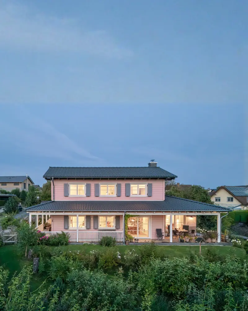 Rückansicht eines rosa Holzhauses mit breiter überdachter Veranda, großen Fenstern, Garten und warm beleuchteten Wohnräumen in der Dämmerung.
