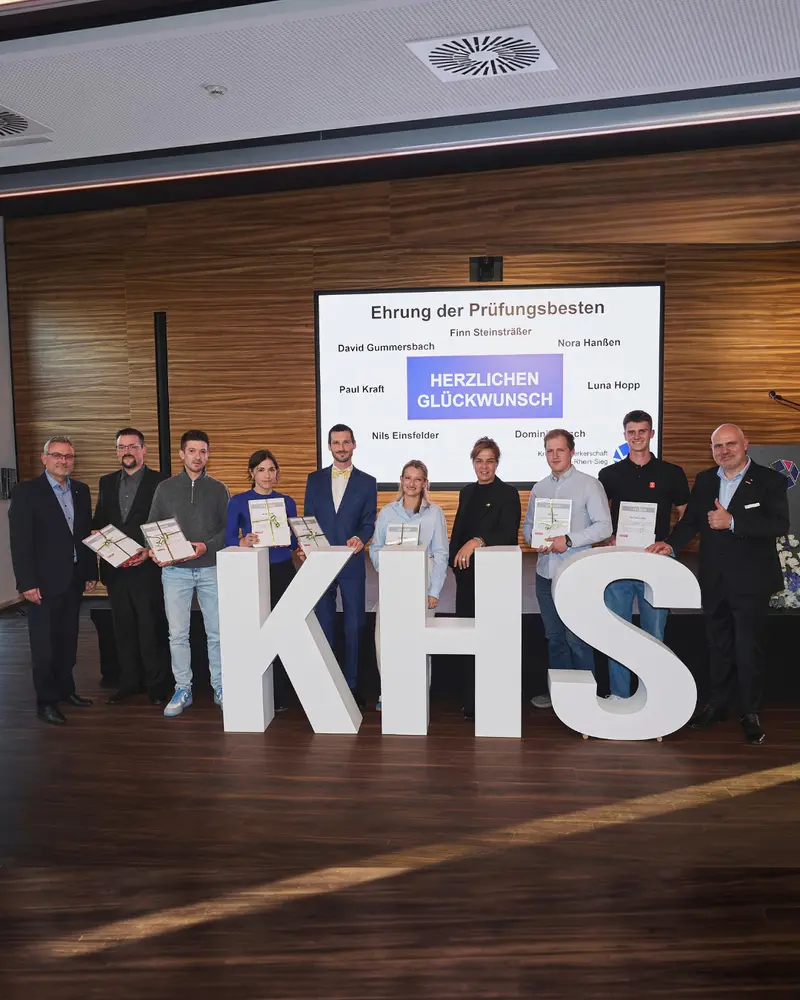 Gruppenaufnahme auf der Bühne mit mehreren ausgezeichneten Personen und Begleitpersonen. Einige halten Urkunden. Im Vordergrund stehen große weiße Buchstaben „KHS“. Auf der Leinwand sind „Ehrung der Prüfungsbesten“, mehrere Namen und „Herzlichen Glückwunsch“ zu lesen.