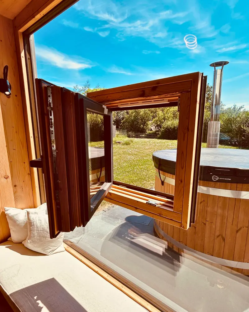 Innenansicht eines Holzhauses mit geöffnetem kleinen Fenster, das in ein großes Glasfenster integriert ist, mit Blick in den Garten