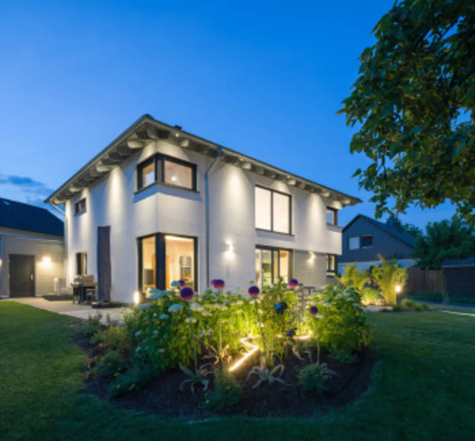 Modernes Stommel-Haus-Einfamilienhaus in der Abenddämmerung mit beleuchteten Fenstern und Blumenbeet im Vordergrund.