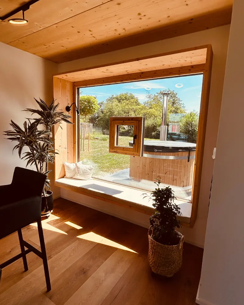 Innenraum eines Holzhauses mit großer Fensterfront, Sitzbank und integriertem kleinen Fenster als zentrales Gestaltungselement