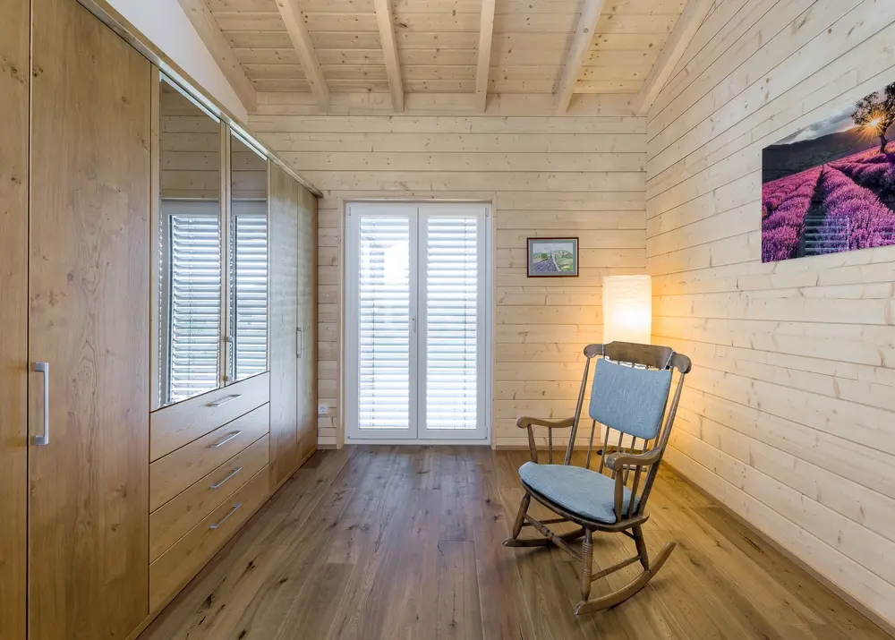 Ein mit Holz ausgekleideter Ankleideraum eines kleinen Holzhauses für Senioren. Ein großer Holzschrank steht zur Linken, ein Schaukelstuhl mitten im Raum.