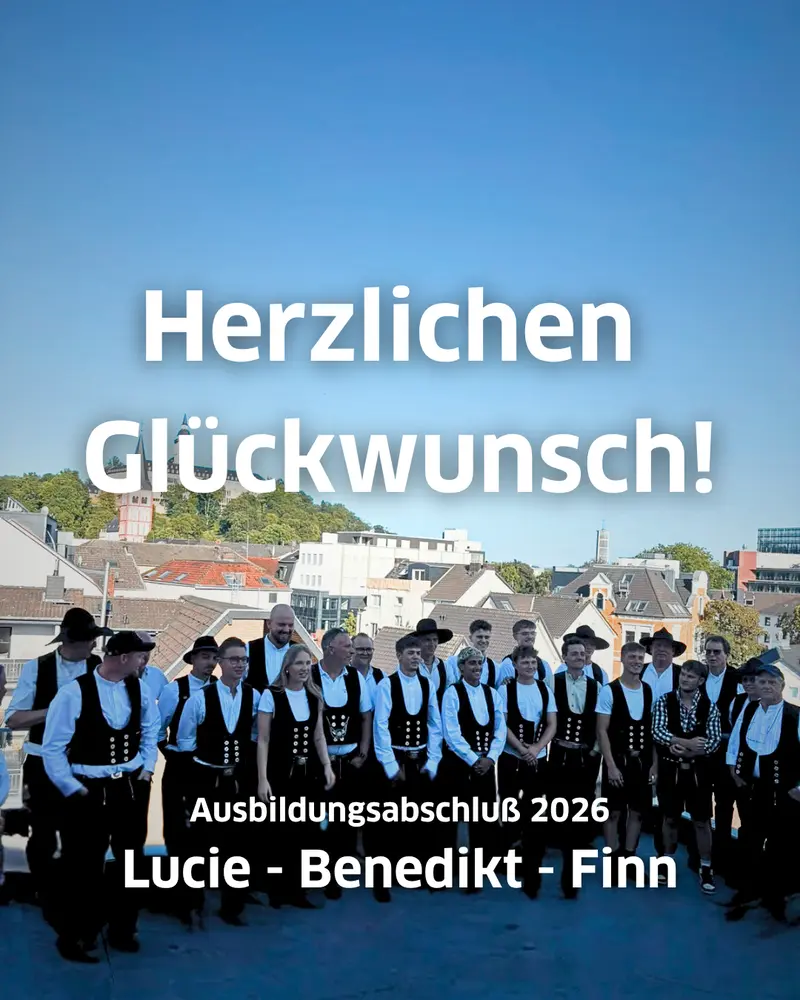 Gruppenfoto mehrerer Zimmerer nach bestandener Gesellenprüfung 2026, vorne drei Auszubildende Lucie, Benedikt und Finn, im Hintergrund Stadtansicht mit Gebäuden und blauem Himmel, Schriftzug „Herzlichen Glückwunsch“ sichtbar