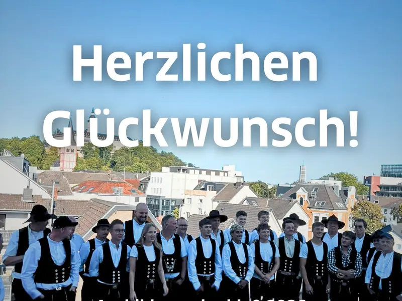 Gruppenfoto mehrerer Zimmerer nach bestandener Gesellenprüfung 2026, vorne drei Auszubildende Lucie, Benedikt und Finn, im Hintergrund Stadtansicht mit Gebäuden und blauem Himmel, Schriftzug „Herzlichen Glückwunsch“ sichtbar