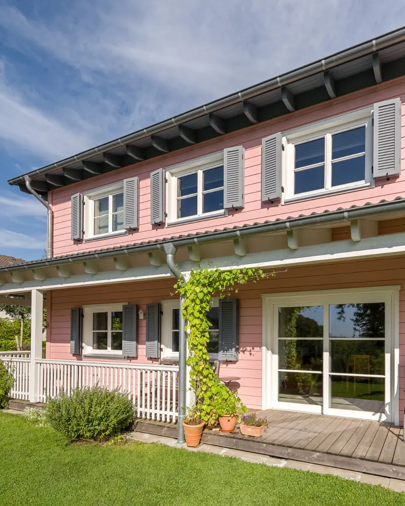 Detail einer rosa Holzfassade mit weißen Fensterrahmen, grauen Fensterläden, überdachter Terrasse, rankender Pflanze und Rasenfläche.