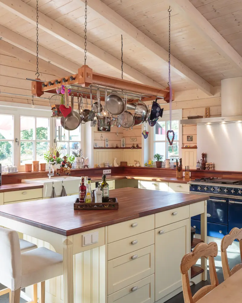 Helle Küche in einem Holzhaus mit Holzdecke, Kochinsel, großen Fenstern, cremefarbenen Fronten und Arbeitsplatten aus Holz.