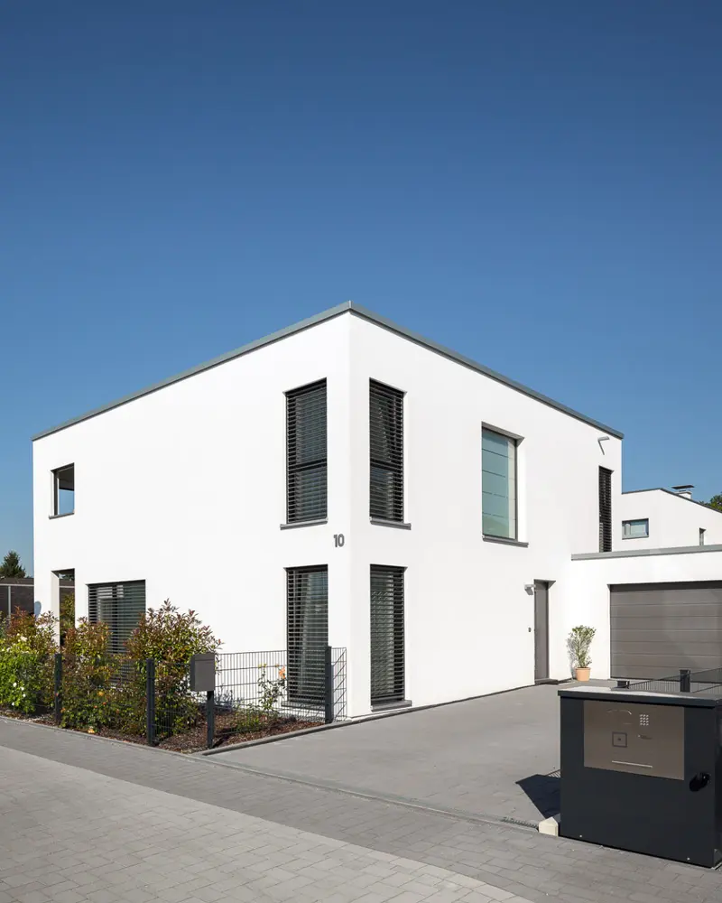 Modernes Einfamilienhaus in klarer Architektur mit Garage, gepflasterter Zufahrt, Einfriedung und angelegten Außenbereichen vor blauem Himmel.