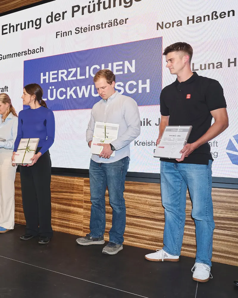 Mehrere ausgezeichnete Personen stehen mit Urkunden auf einer Bühne. Eine Person auf der rechten Seite trägt ein schwarzes Poloshirt und hält eine gerahmte Urkunde. Auf der Leinwand stehen „Ehrung der Prüfungsbesten“, „Finn Steinsträßer“ und „Herzlichen Glückwunsch“.