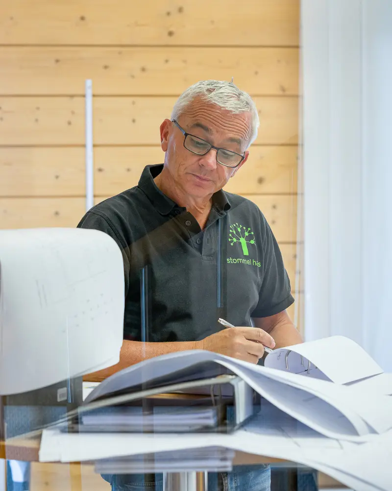 Eine Person arbeitet konzentriert an Bauzeichnungen und überprüft Planunterlagen auf einem Tisch mit Ordnern und technischen Zeichnungen