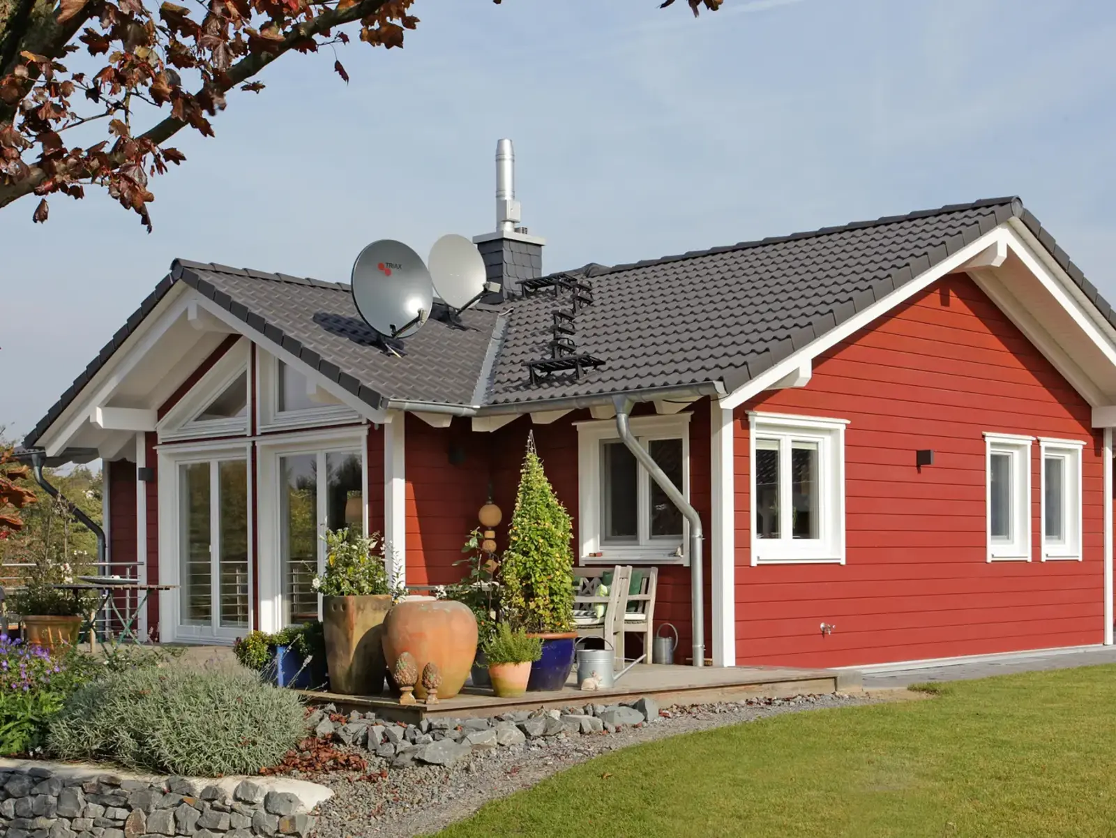 Eingeschossiger Holzbungalow mit roter Holzfassade, weißen Fensterrahmen und überdachter Terrasse mit Pflanzkübeln.