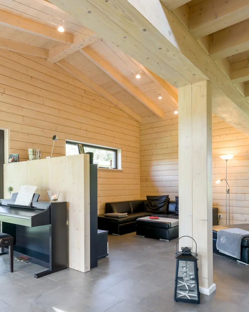 Innenraum eines Massivholzhauses mit sichtbaren Holzwänden, kräftigen Holzbalken, heller Holzdecke, Sitzbereich und Klavier.
